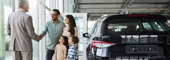 Family standing in an auto dealership smiling looking at a sales person. The sales person is shaking hands with the dad. 