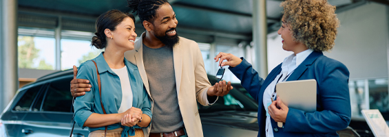 Couple stand together at a auto dealer smiling. Sale person looking the couple in front of a new vehicle.