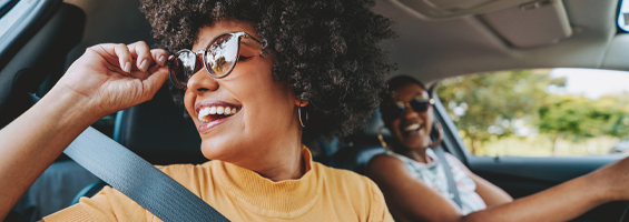 Two women in a car. One looking out the window smiling and other smiling while looking forward. 