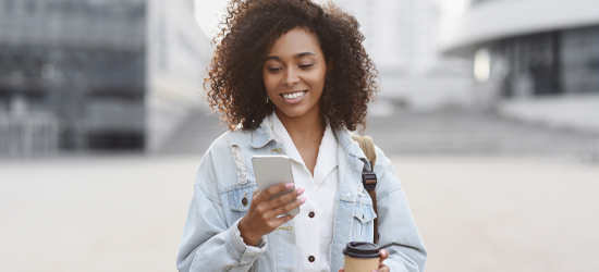 Women standing outside holding a cellphone and coffee. She is looking at the her phone smiling