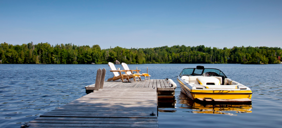 Dock we two chairs in middle of photos surrounded by water and green trees in the background. A yellow boat attached to the dock.